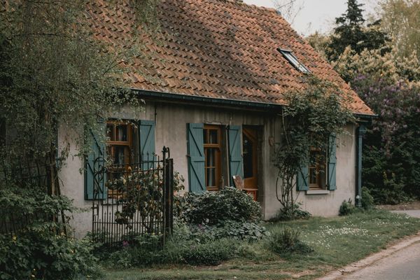 An old tiled-roof cottage with faded blue shutters sits behind a wrought iron garden gate and an overgrown front garden.