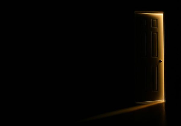 A panelled door stands slightly ajar in a pitch dark room, warm light spilling around its edges and across the floor.
