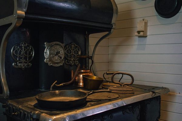 An antique cast iron kitchen range with ornate nickel trim holding a copper kettle and a brass cup and a skillet and old flat irons.