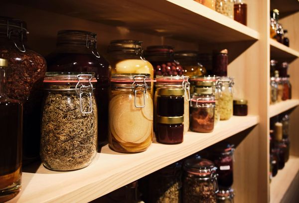 Clip-top jars of dried herbs, preserved fruit and jam line warm wooden pantry shelves in soft low light.