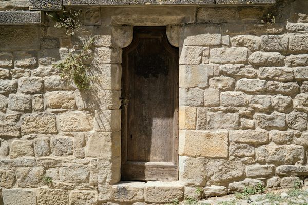 A narrow weathered wooden door set in an old sandstone wall above a worn stone threshold with weeds growing at its edges.