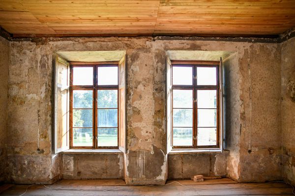 An emptied old room with bare plaster walls, a pine board ceiling and two tall wooden windows letting in green garden light.