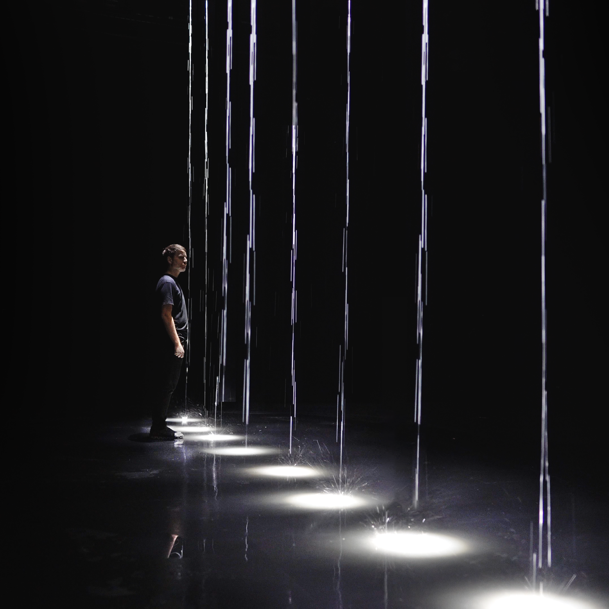 Un homme sur scène, faisant face à plusieurs colonnes de gouttes d'eau qui s'abattent sur le plancher, accompagnées de faisceaux lumineux.