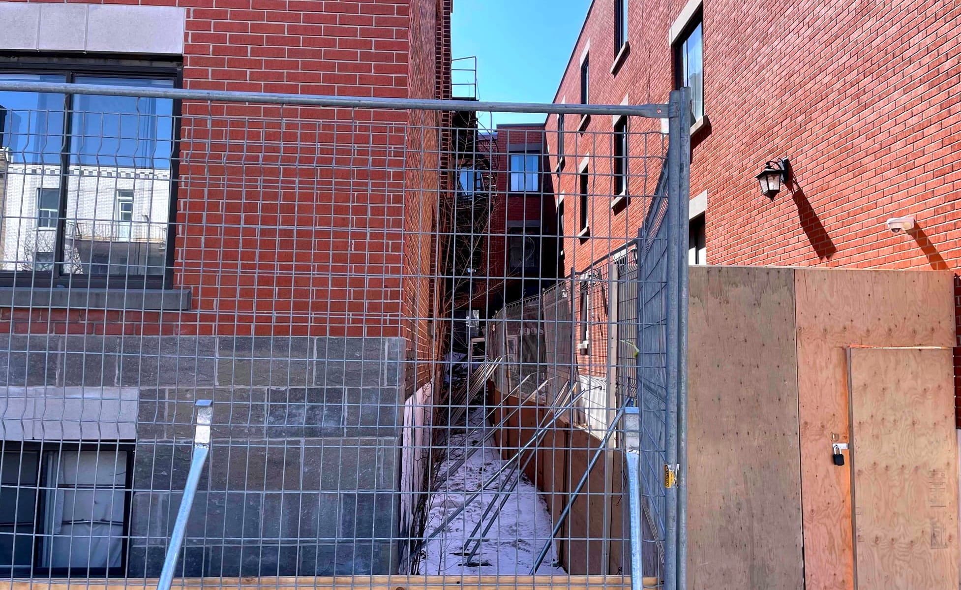 On voit un grillage et des panneaux de bois qui bloquent l'entrée d'un édifice en briques.