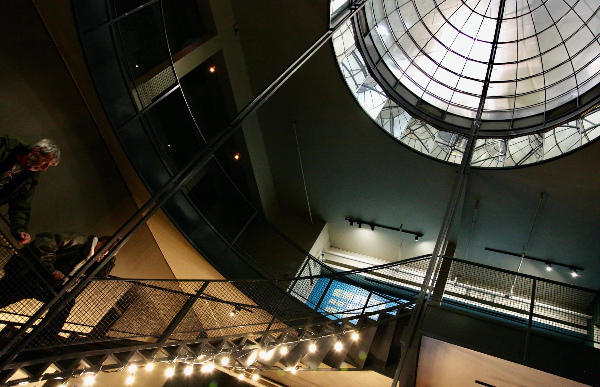 On voit deux personnes dans un escalier bordé de lumières et tout en haut un dôme avec vitres et miroirs.