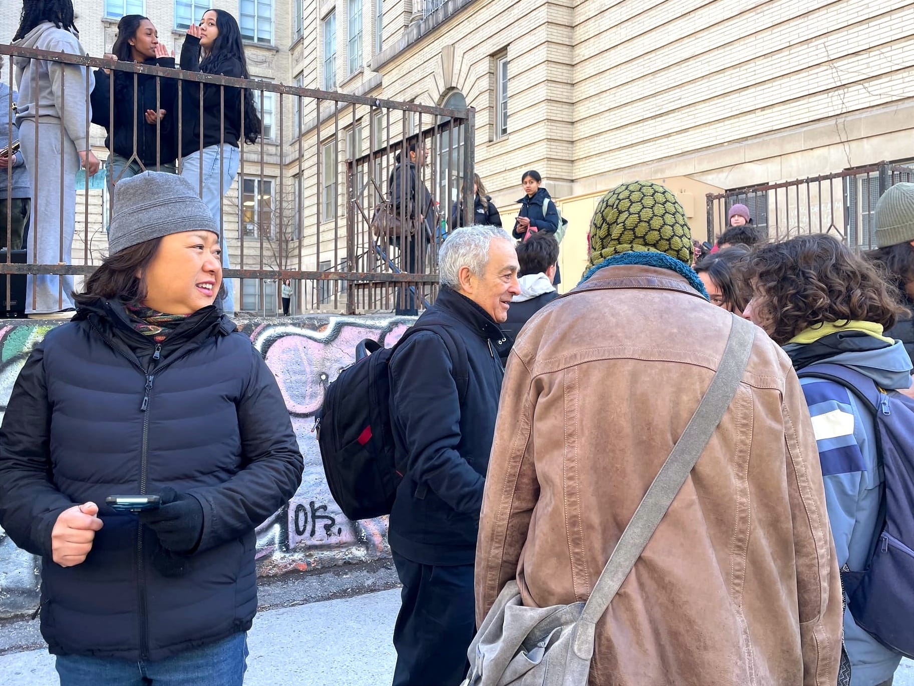 On voit une enseignante à la gauche et Raymond à sa gauche, discutant avec un petit cercle de personnes.