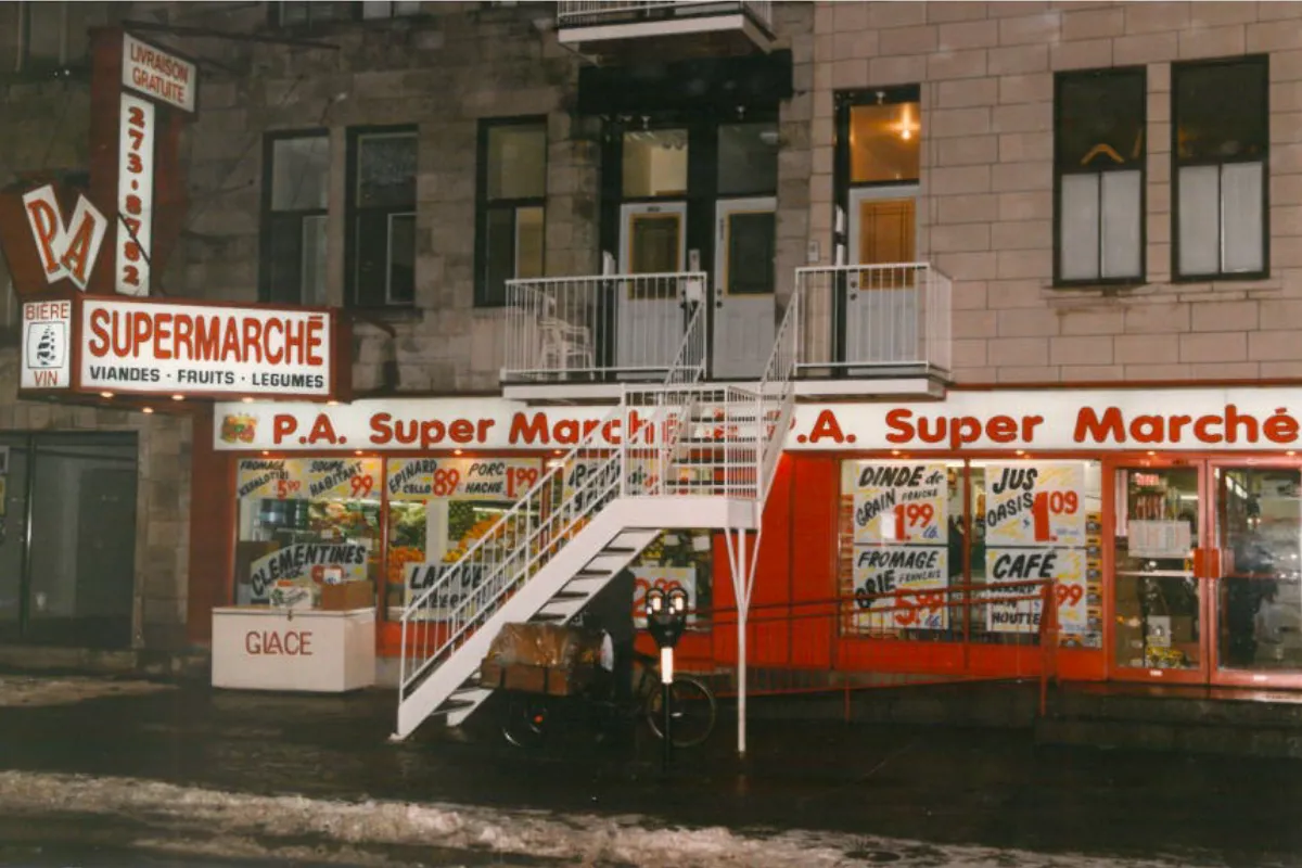 On voit l'ancienne devanture de l'épicerie avec le prix des aliments affiché dans les fenêtres.