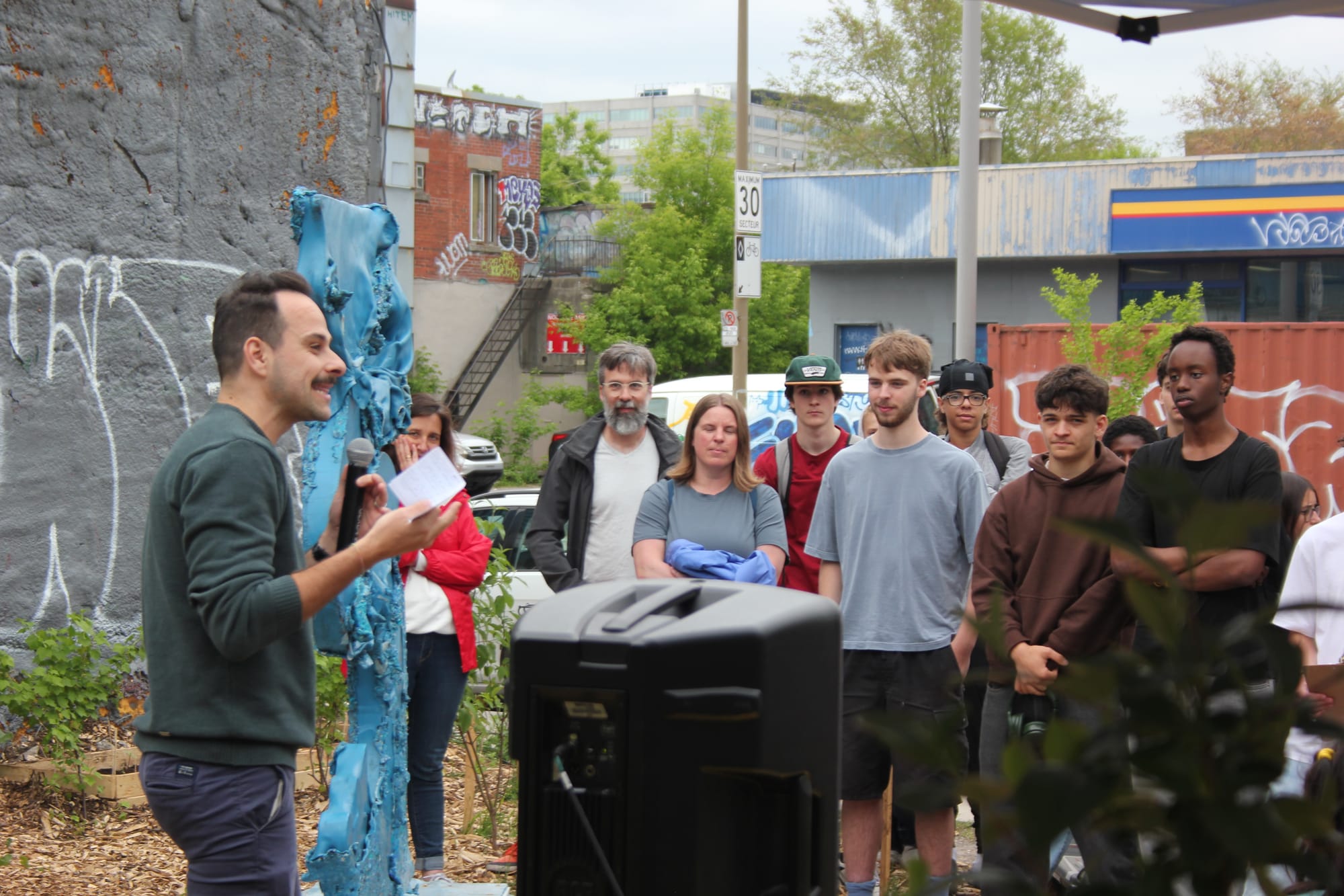 M. Turgeon-Côté s'exprime au microphone, à côté de la sculpture avec une dizaine de personnes qui l'écoutent en arrière-plan.