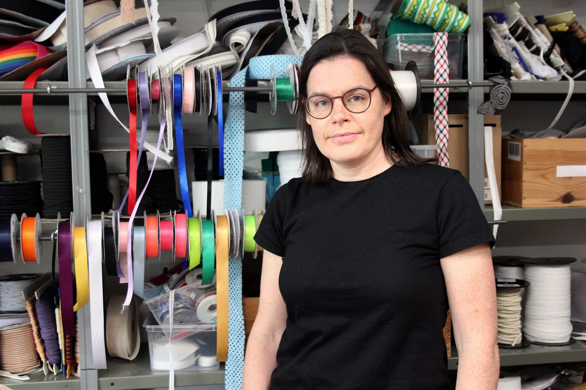 Mme Lussier se tient debout devant un inventaire de rubans et autres accessoires de couture.