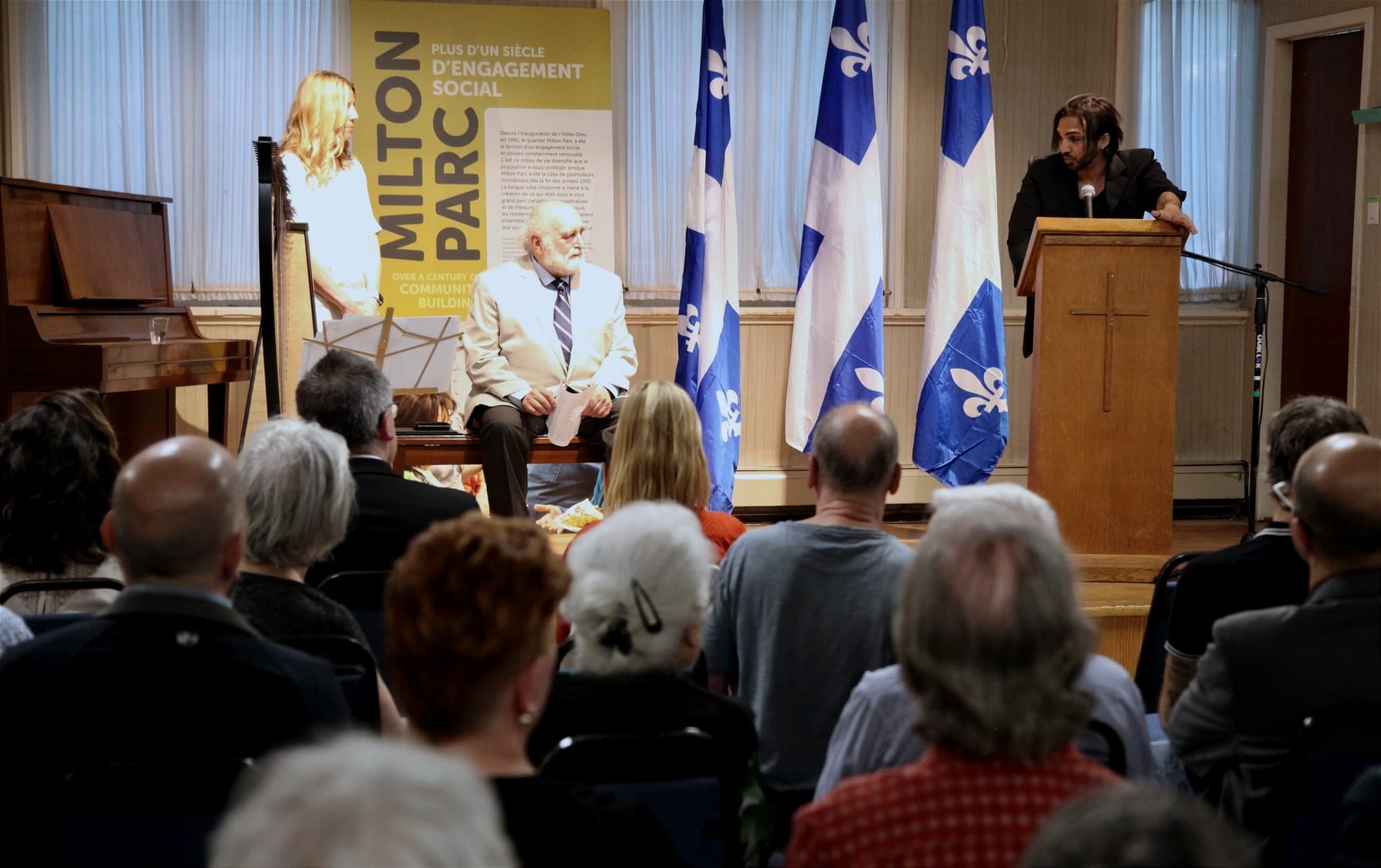 Du milieu de la salle on voit M. Aliskandaria au podium, M. Phaneuf l'écoutant, assis sur scène, et Mme Maccarone, debout, à côté de ce dernier.