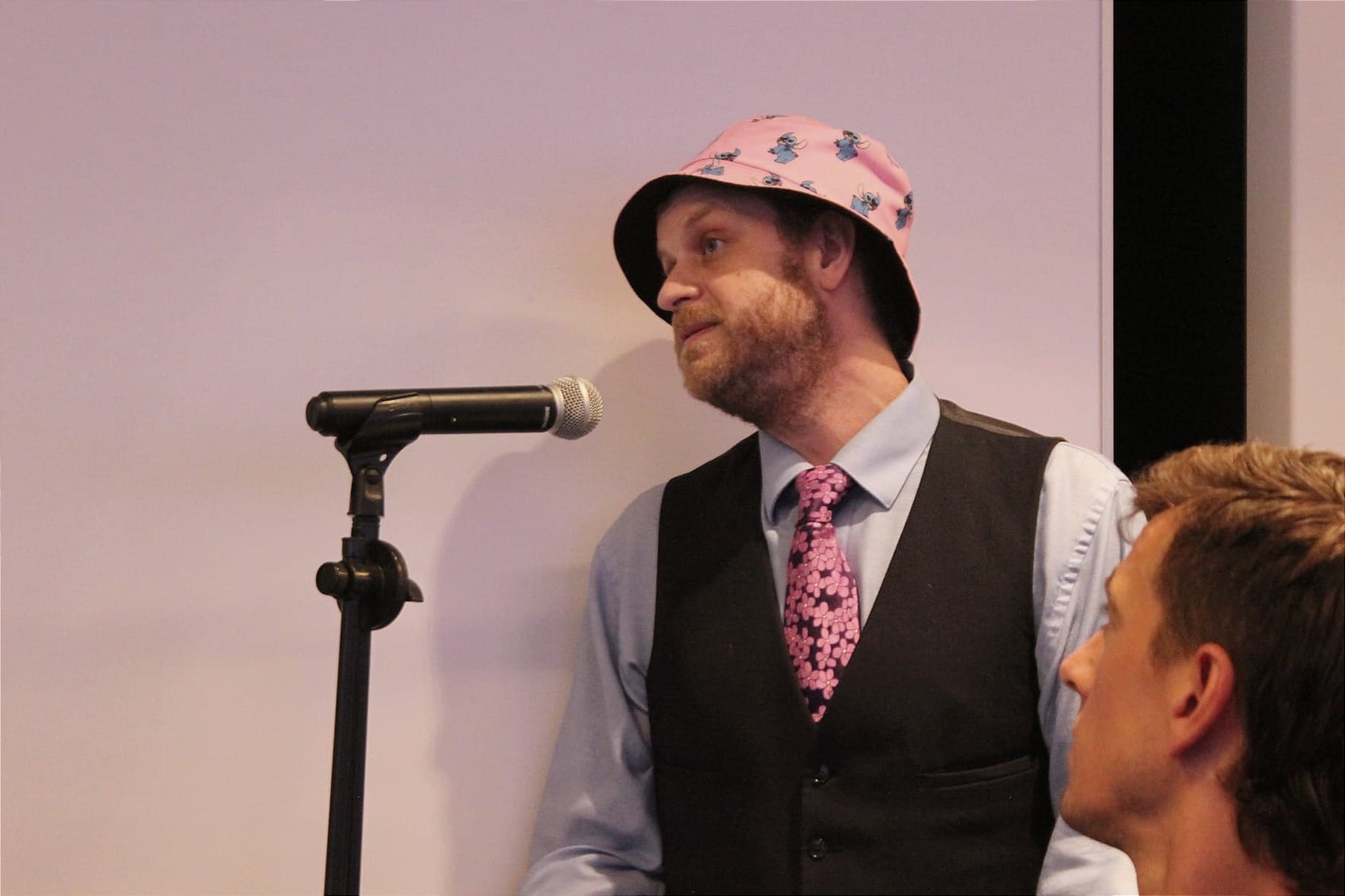 M. Corelli devant le micro, vêtu d'une veste noire, d'une cravate et d'un chapeau de pêche rose avec des personnages. Son regard est dirigé vers les élus. 