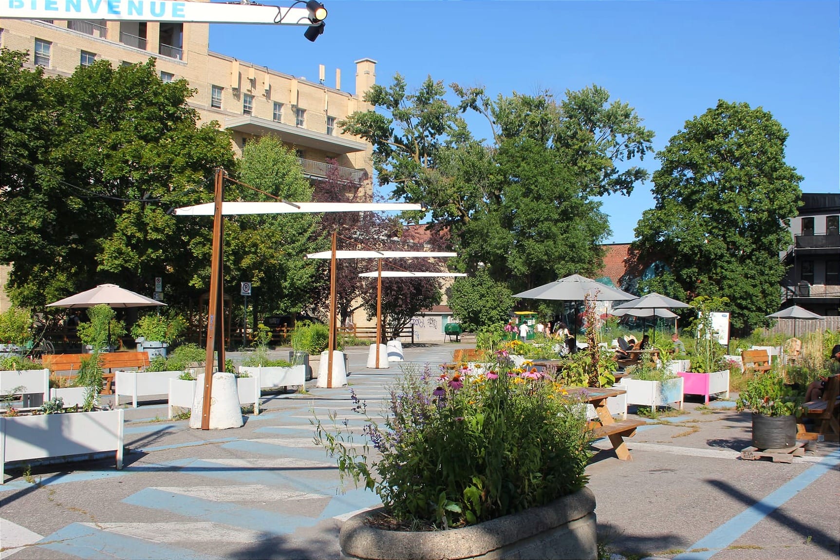 Perspective sur le terrain, regardant vers le sud-est. On voit plusieurs bacs, des parasols, tables et bancs, ainsi que le Centre de services communautaires du Monastère en arrière-plan. 
