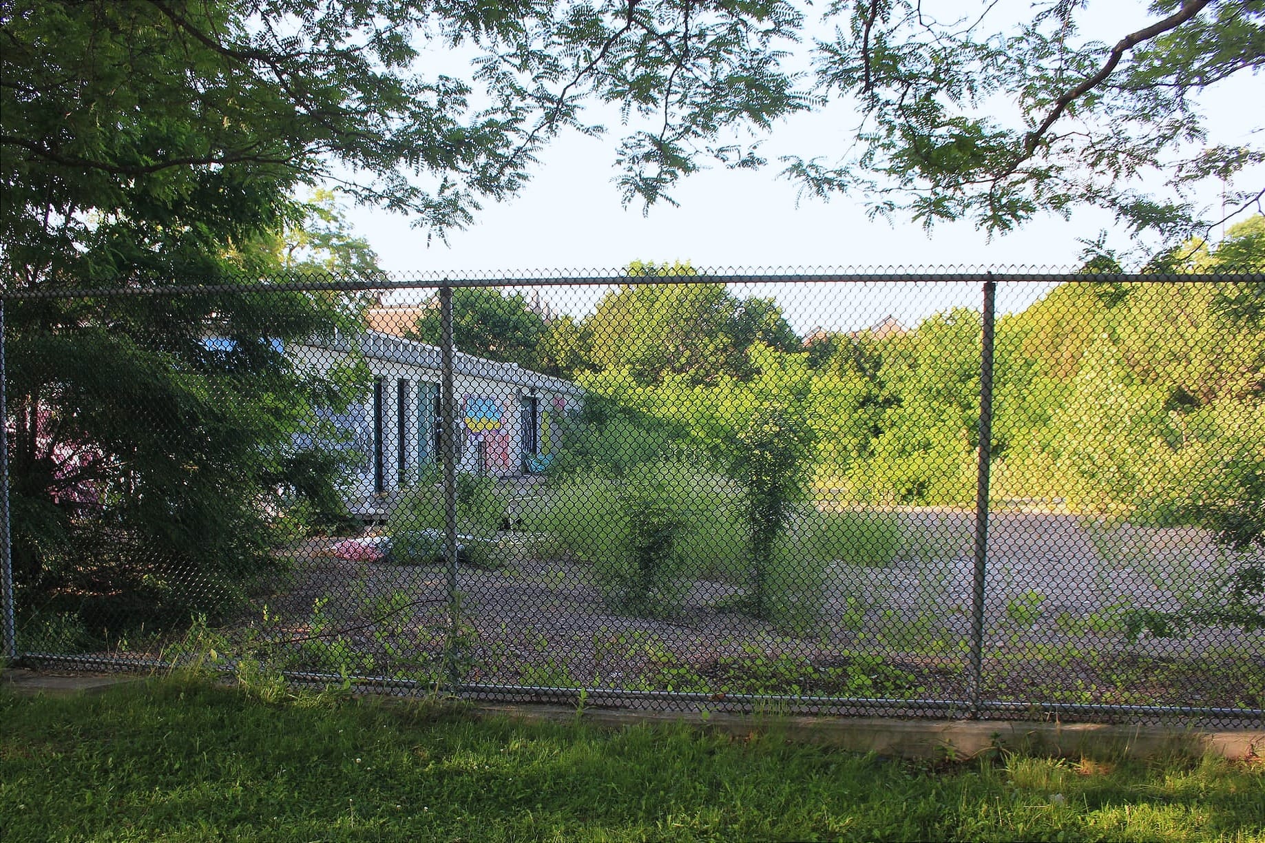 Au travers d'une clôture, on peut voir l'ancien chalet, mais la piscine en soi a disparue et laissé place à de la terre sèche et de la végétation sauvage.