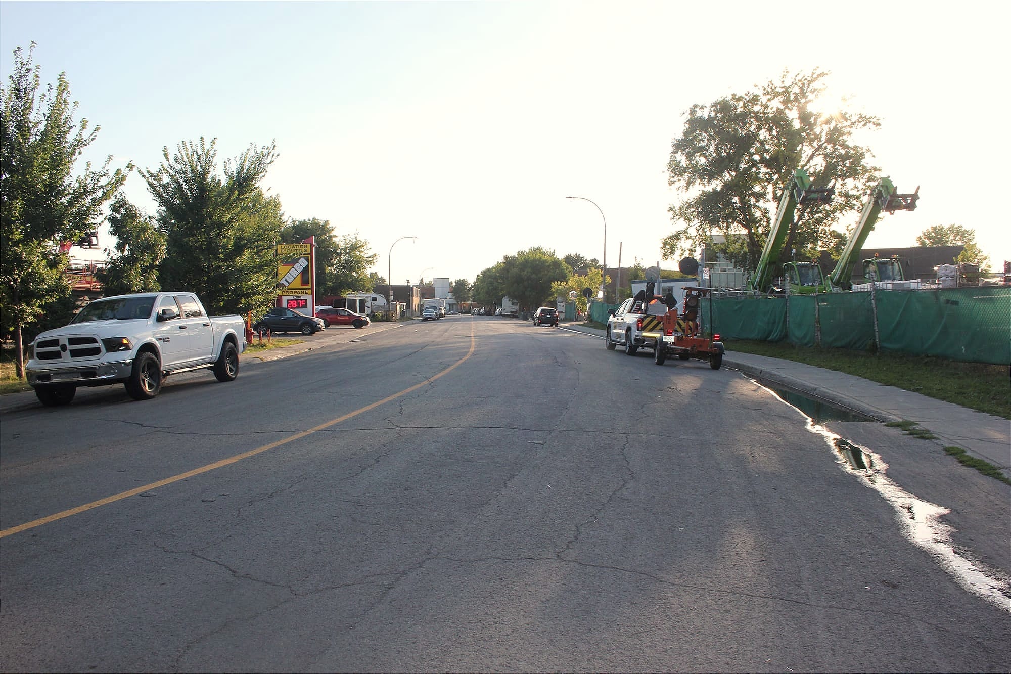 On voit une vaste rue avec peu de véhicules garés, et des véhicules de constructions qui dépassent des clôtures de grandes cours industrielles.