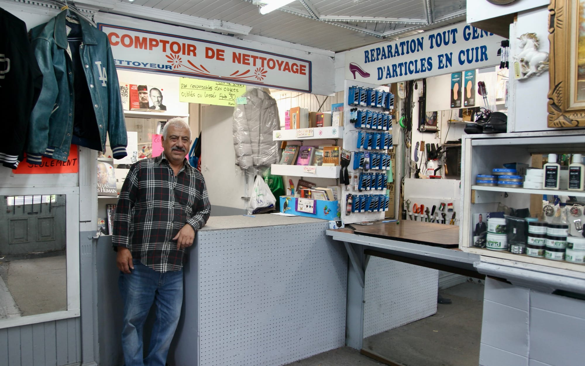 Un homme en chemise et jeans, avec des cheveux blancs, pose devant le comptoir de sa boutique.