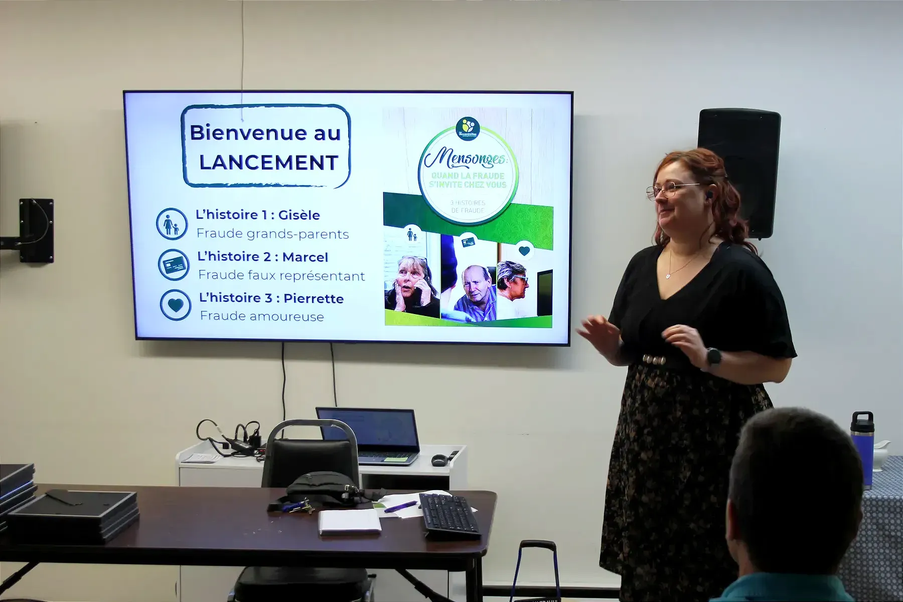 Mme Ostiguy se tient à proximité d'un écran et s'adresse à la petite foule. 
