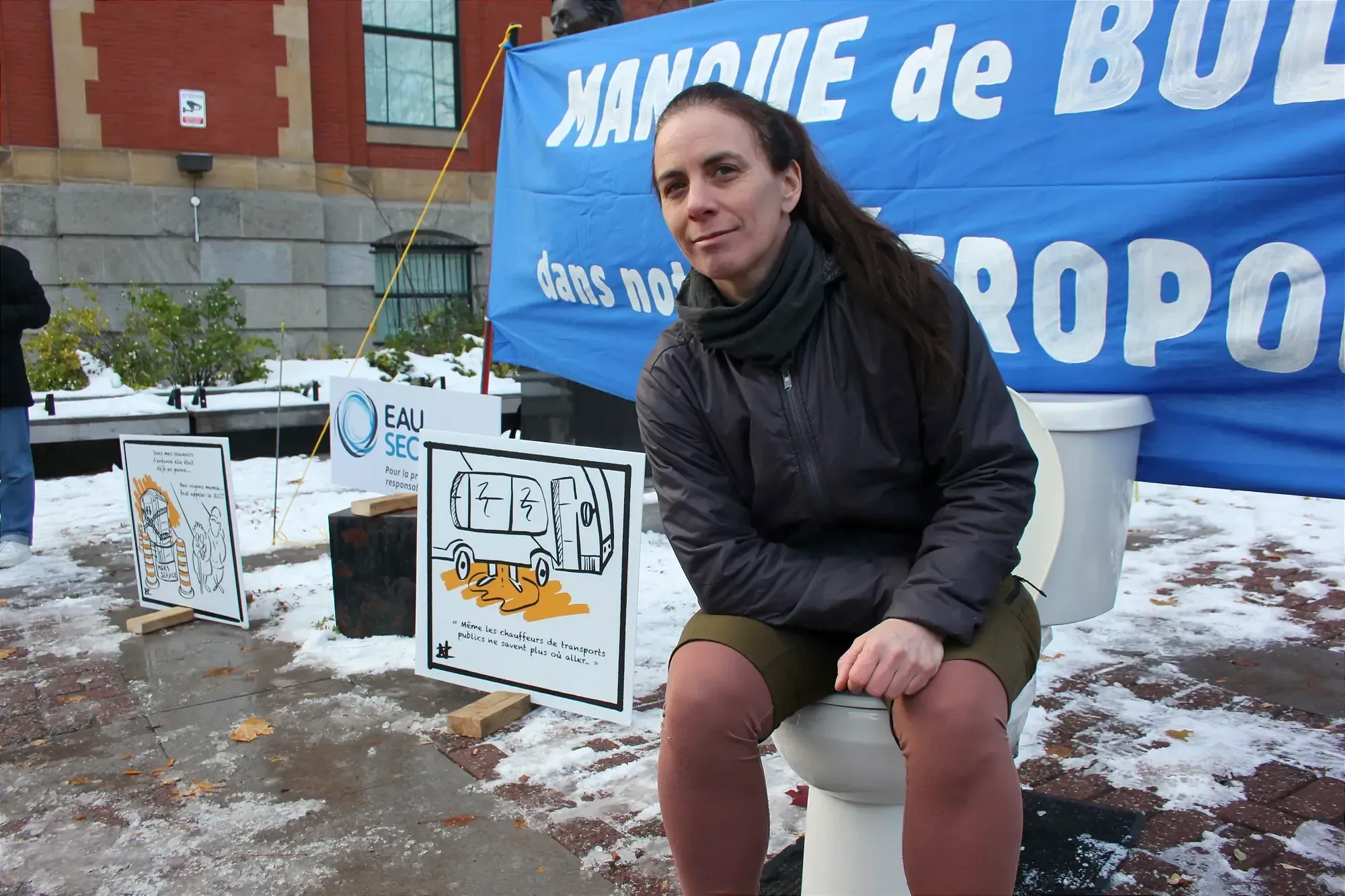 Mme Pétrin pose assise sur la cuvette, devant la banderole et autres affiches de la manifestation. 