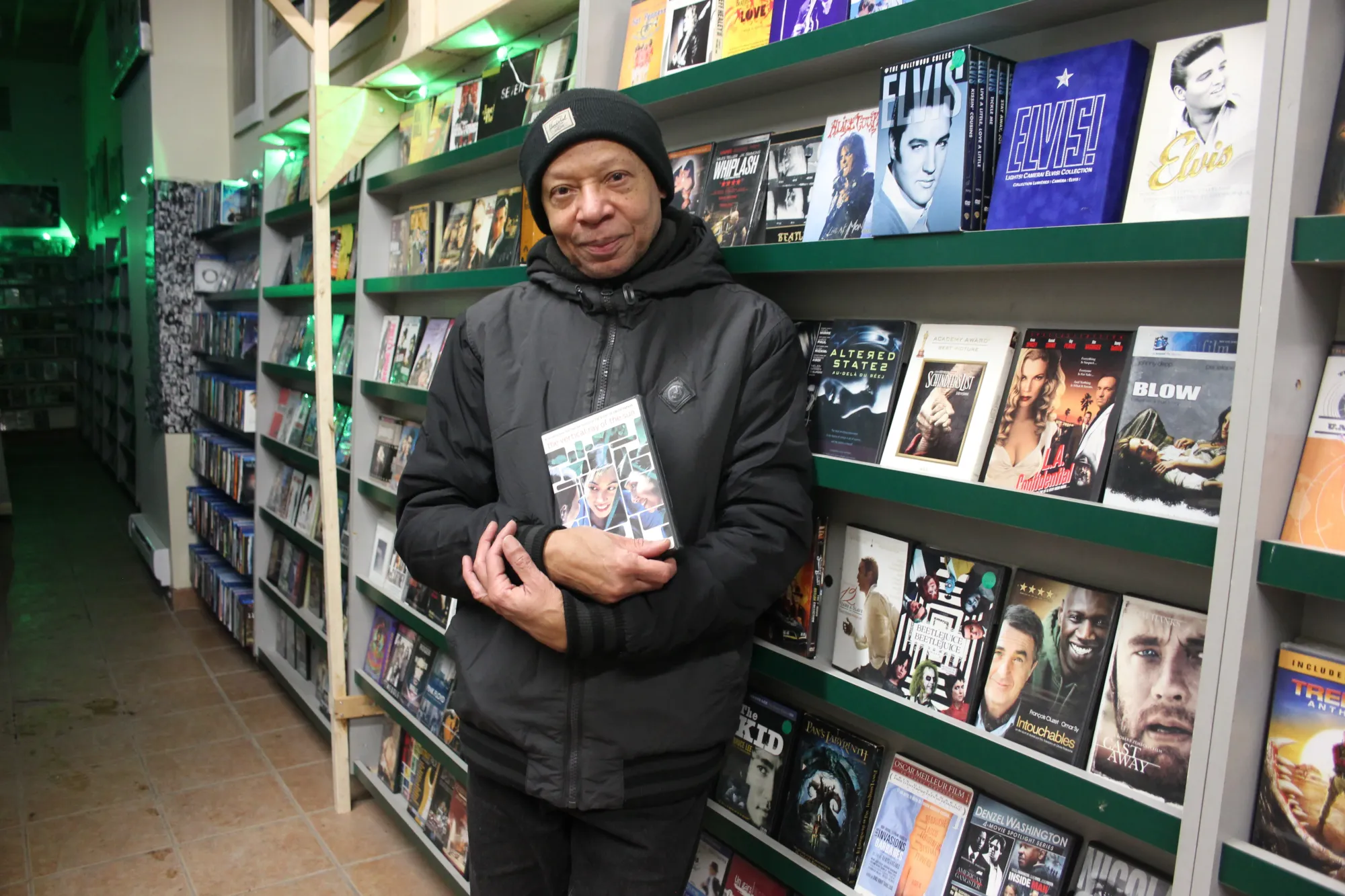 Il porte une tuque, un manteau noir et s'accote sur les étagères.