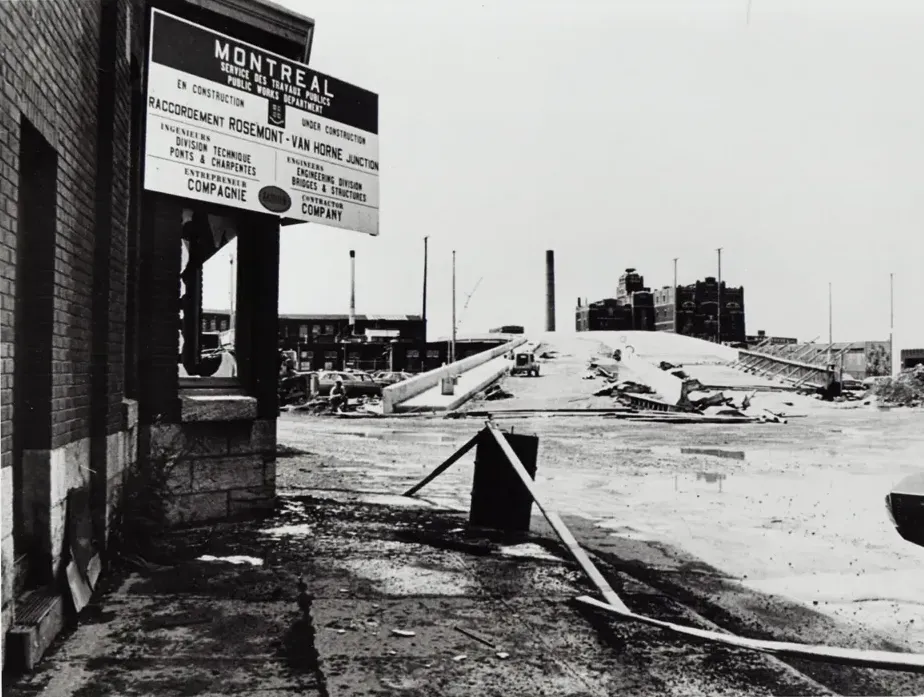 Une photo en noir et blanc du viaduc en construction, en regardant vers l'est. À gauche, on y voit une pancarte des services des travaux publics de Montréal.