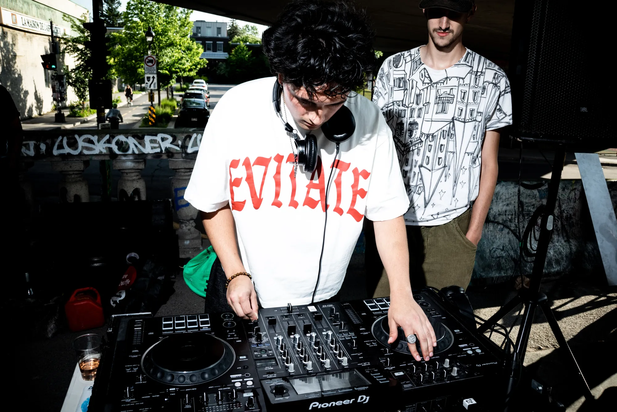 On voit le jeune musicien électronique en action, alors qu'un autre jeune homme derrière lui l'observe, les mains dans les poches.