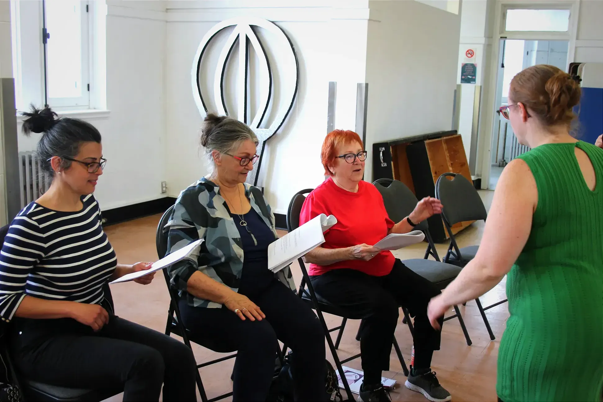 Trois dames sont assises, texte à la main, répétant une portion rythmée de celui-ci, dirigée par une autre femme faisant dos à la caméra.