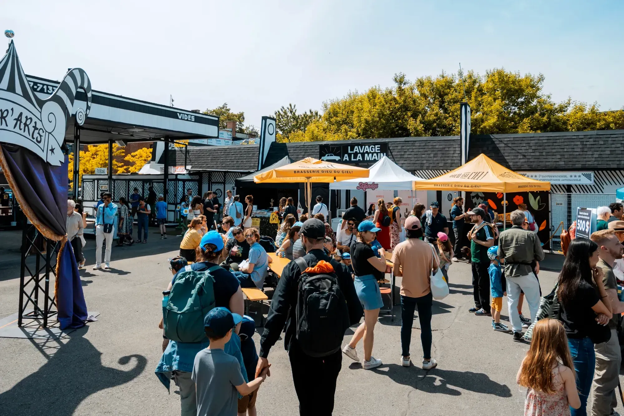 Le site est ensoleillé avec plusieurs personnes debout, explorant les kiosques, ou assises sur des tables de pique-nique.