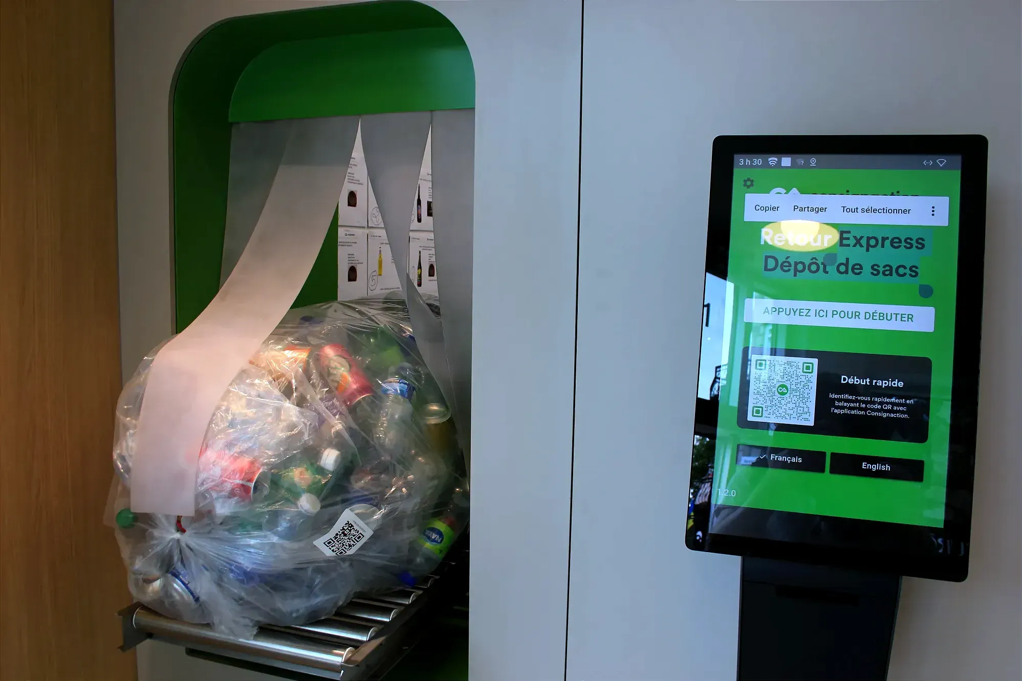 Un sac rempli de bouteilles et de cannettes est placé sur une plateforme roulante à la gauche d'un écran servant au dépôt express de sacs.