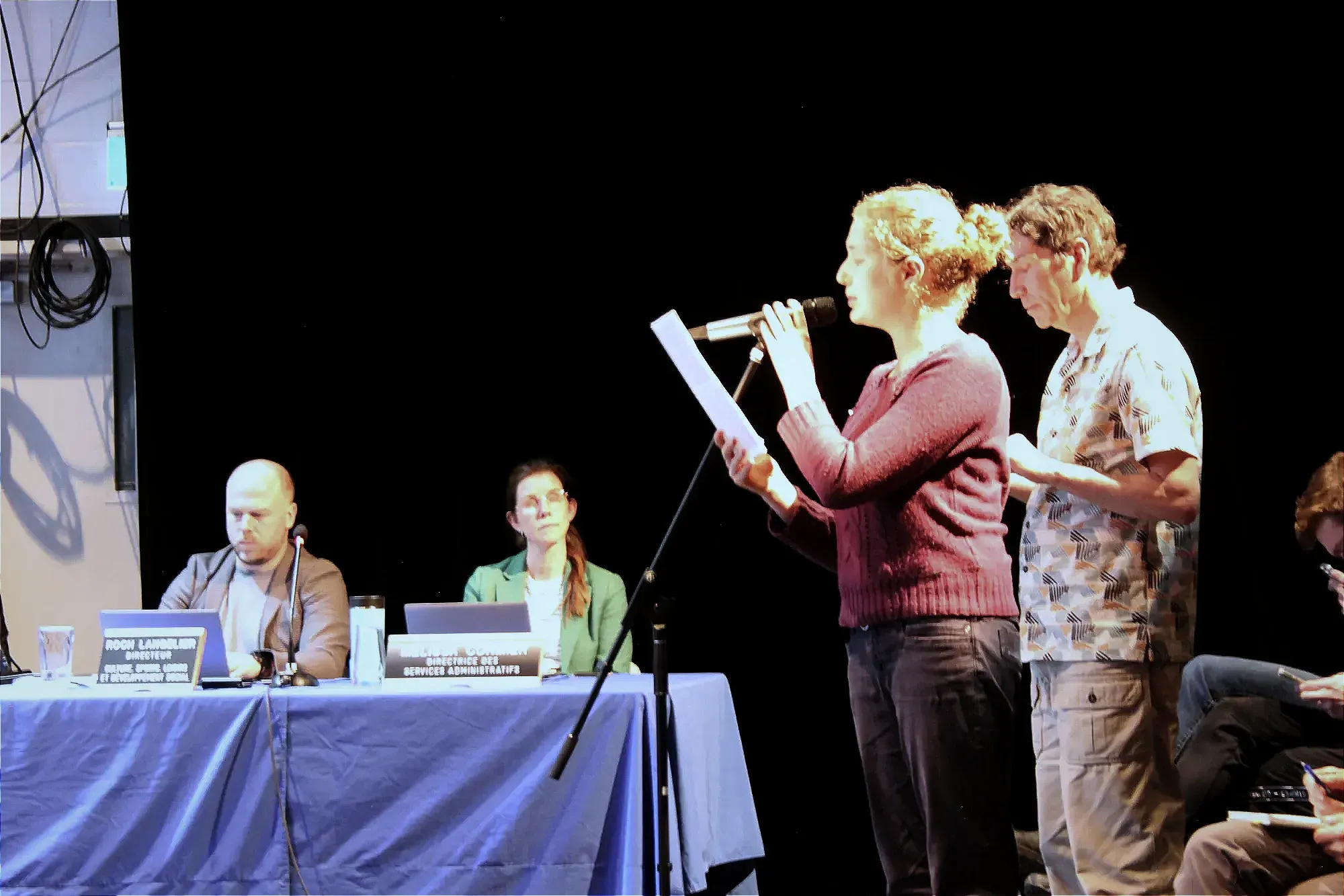 Mme Barneaa est debout, accompagnée d'un homme qui se tient debout derrière elle, alors que les directeurs de l'arrondissement sont à l'écoute en arrière-plan.