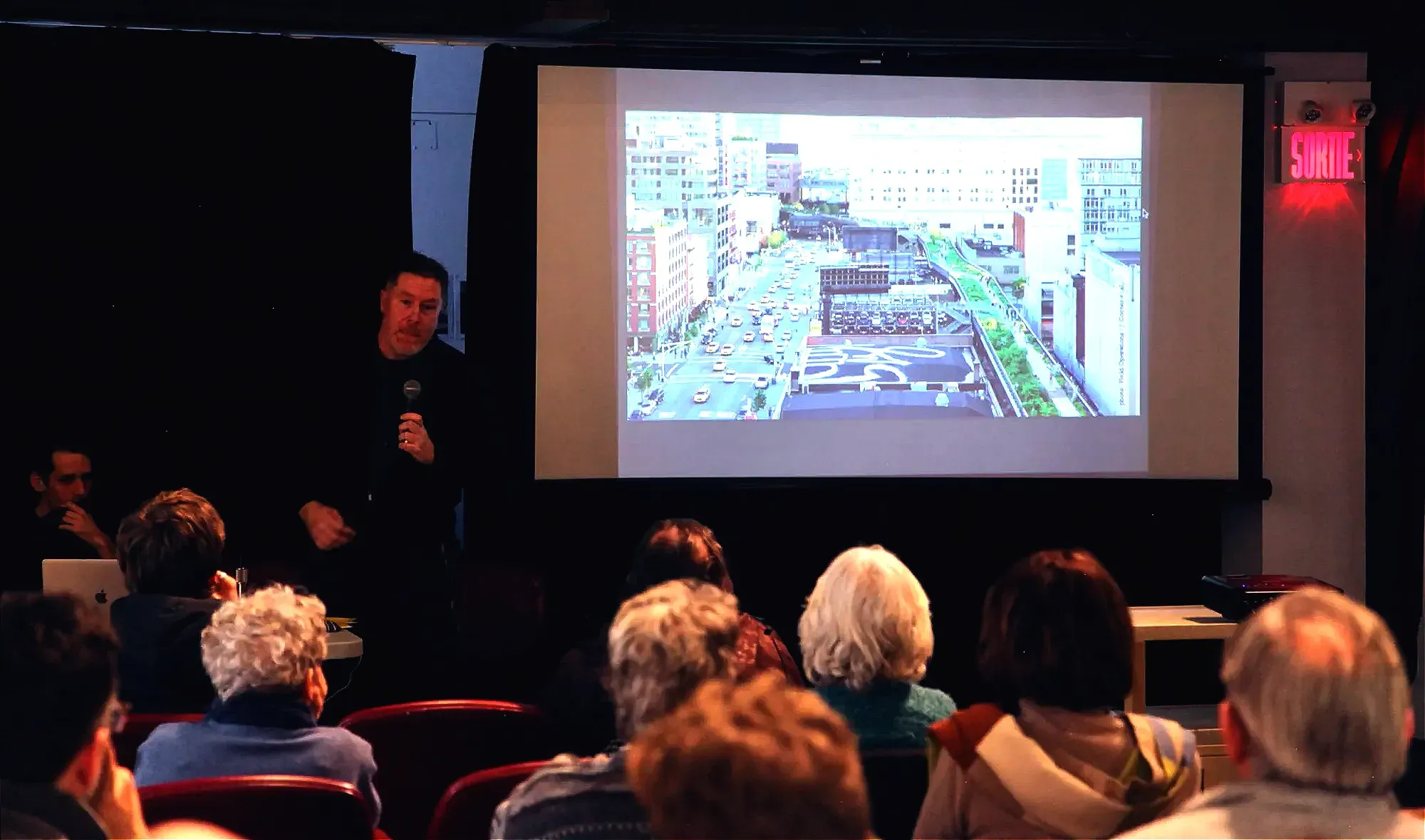 M. Lukas tient un micro devant quelques rangées de spectateurs, alors que la projection d'une image de New York est diffusée sur un écran à ses côté.