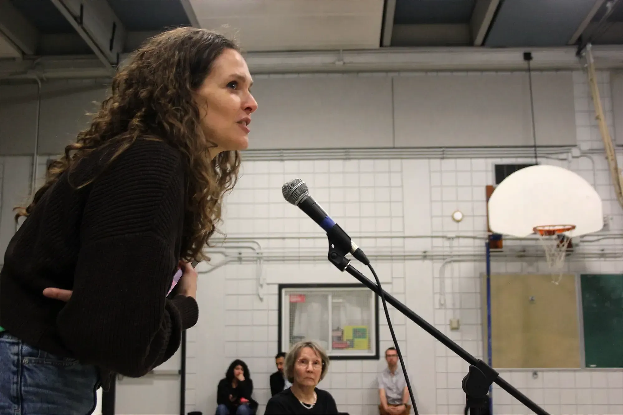 La dame se pence légèrement pour parler dans le microphone, sous le regard de quelques autres personnes présentes dans le gymnase de l'école Saint-Enfant-Jésus.