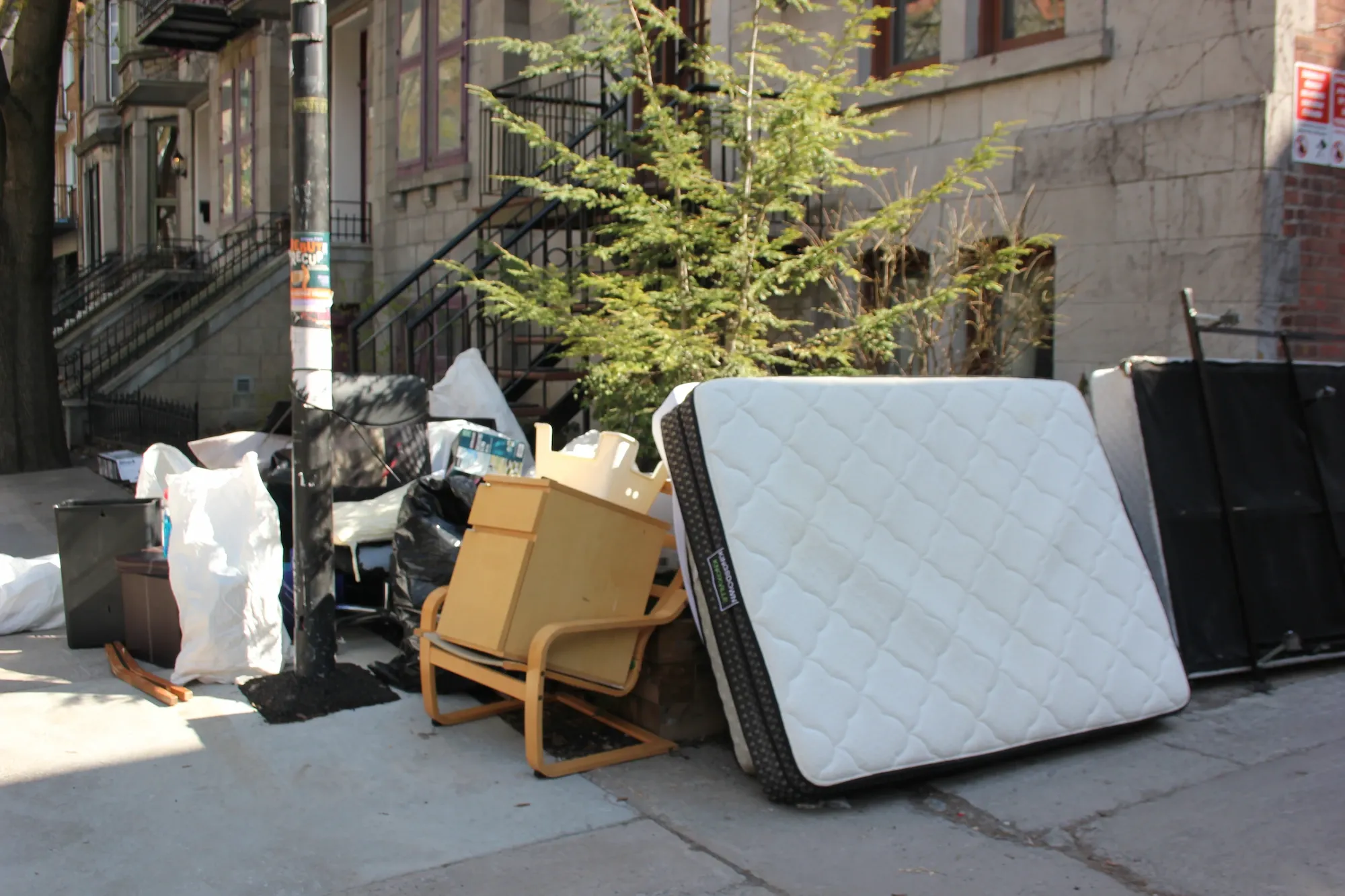 On peut voir un matelas, une chaise, un tabouret, une petite commode et plusieurs sacs.