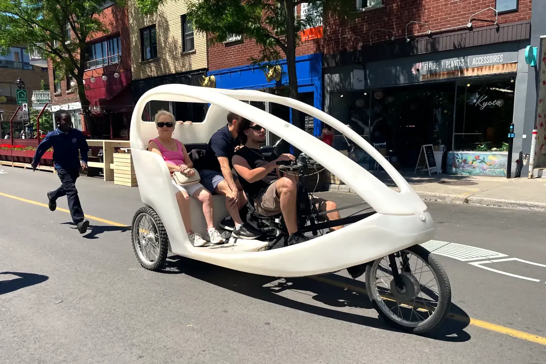 Le véhicule ressemble à un vélo allongé à trois roues, sur lequel une carrosserie en plastique blanc a été ajoutée, qui comprend une banquette arrière pour passagers.