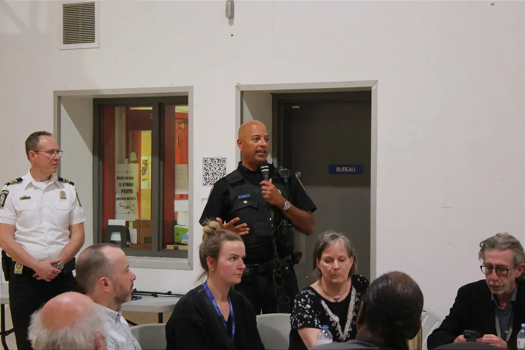 L'agent Dyke s'adresse aux participants en utilisant un micro. On le voit derrière quelques personnes assises à une table, et à côté du commandant Malo.
