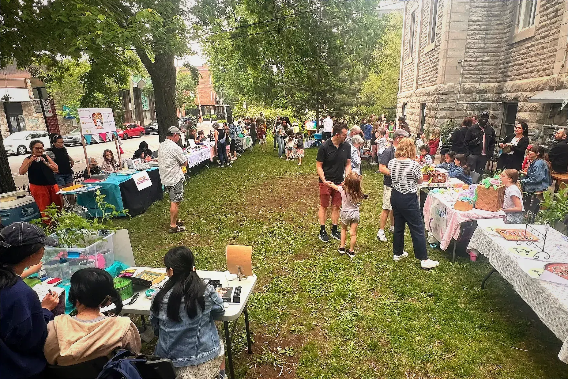 En été, de nombreuses tables avec des produits sont installés sur l'herbe, entourées d'enfants.