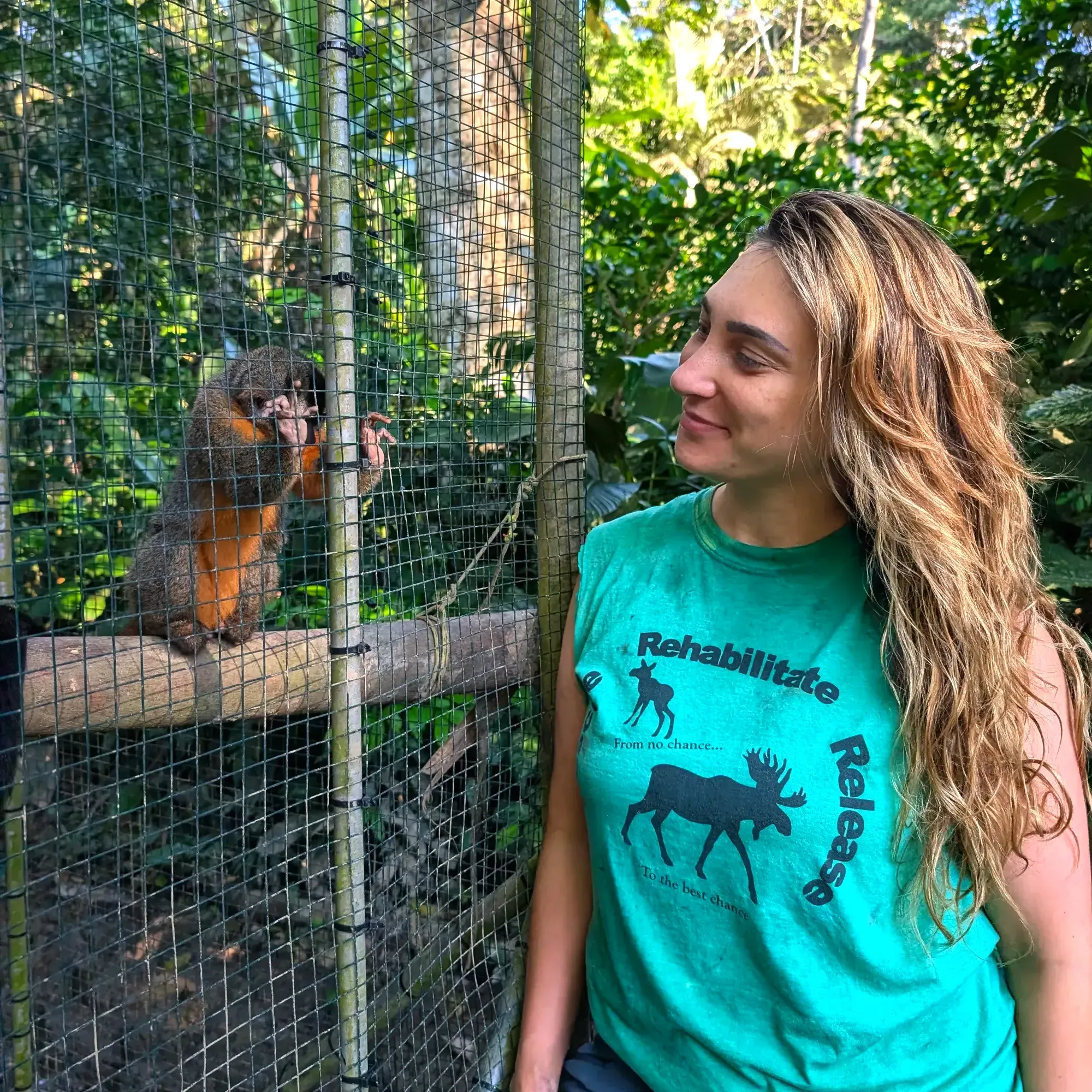 Mme Gerwing, souriante, observe un singe-chouette, à l'intérieur d'une cage située dans ce qui semble être une forêt tropicale. 