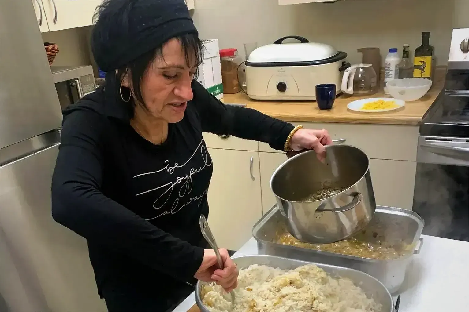 Mme Caserta, casserole dans la main gauche, semble vider des patates pilées dans un large récipient avec un ustensile dans son autre main.