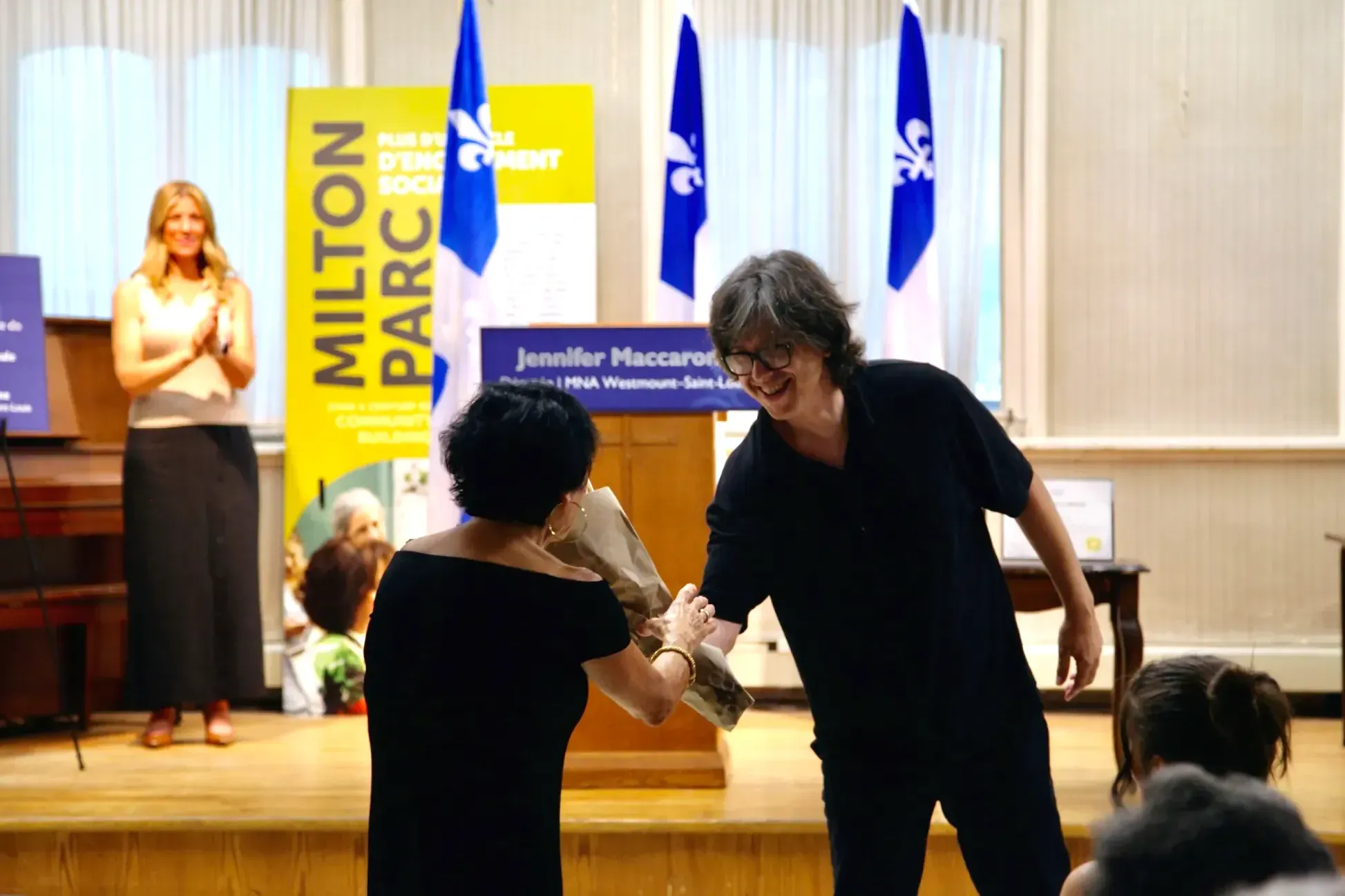 Antonia Caserta, de dos, reçoit un bouquet de fleur. Derrière, sur une petite scène, on peut voir Jennifer Maccarone, éclairée mais floue. 