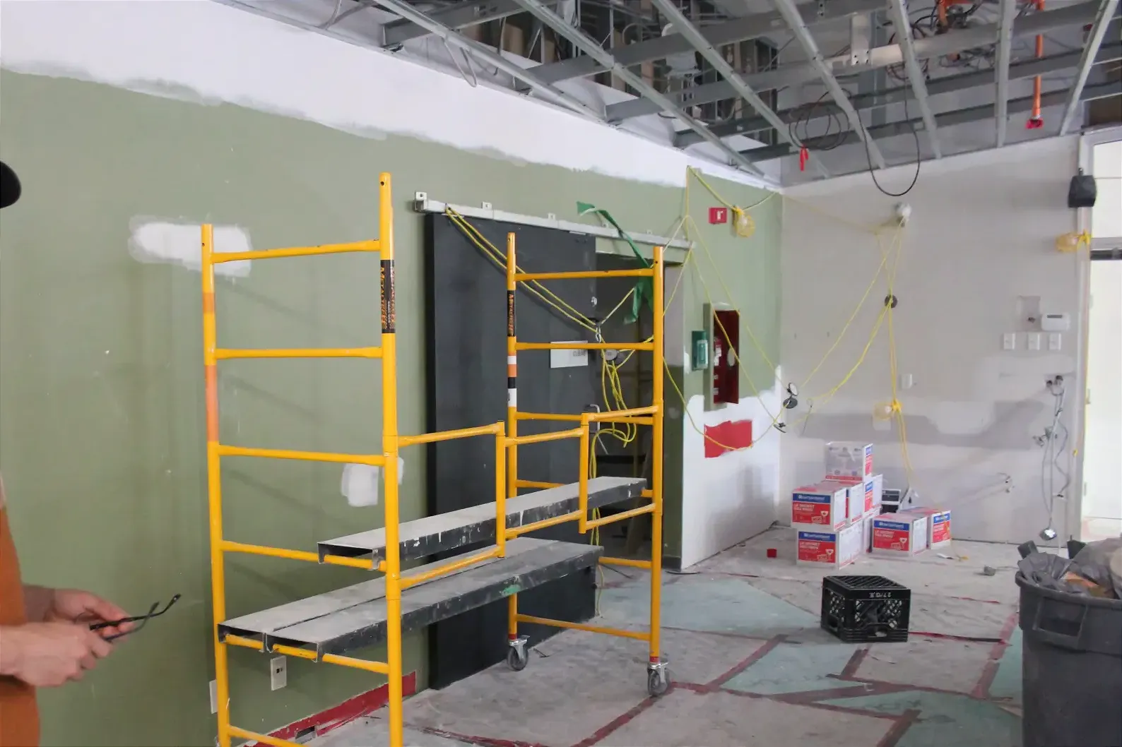 Un échafaudage traîne dans la grand salle du centre de jour. Un grillage métallique est à découvert au plafond et le plancher est encore recouvert de plastique. 