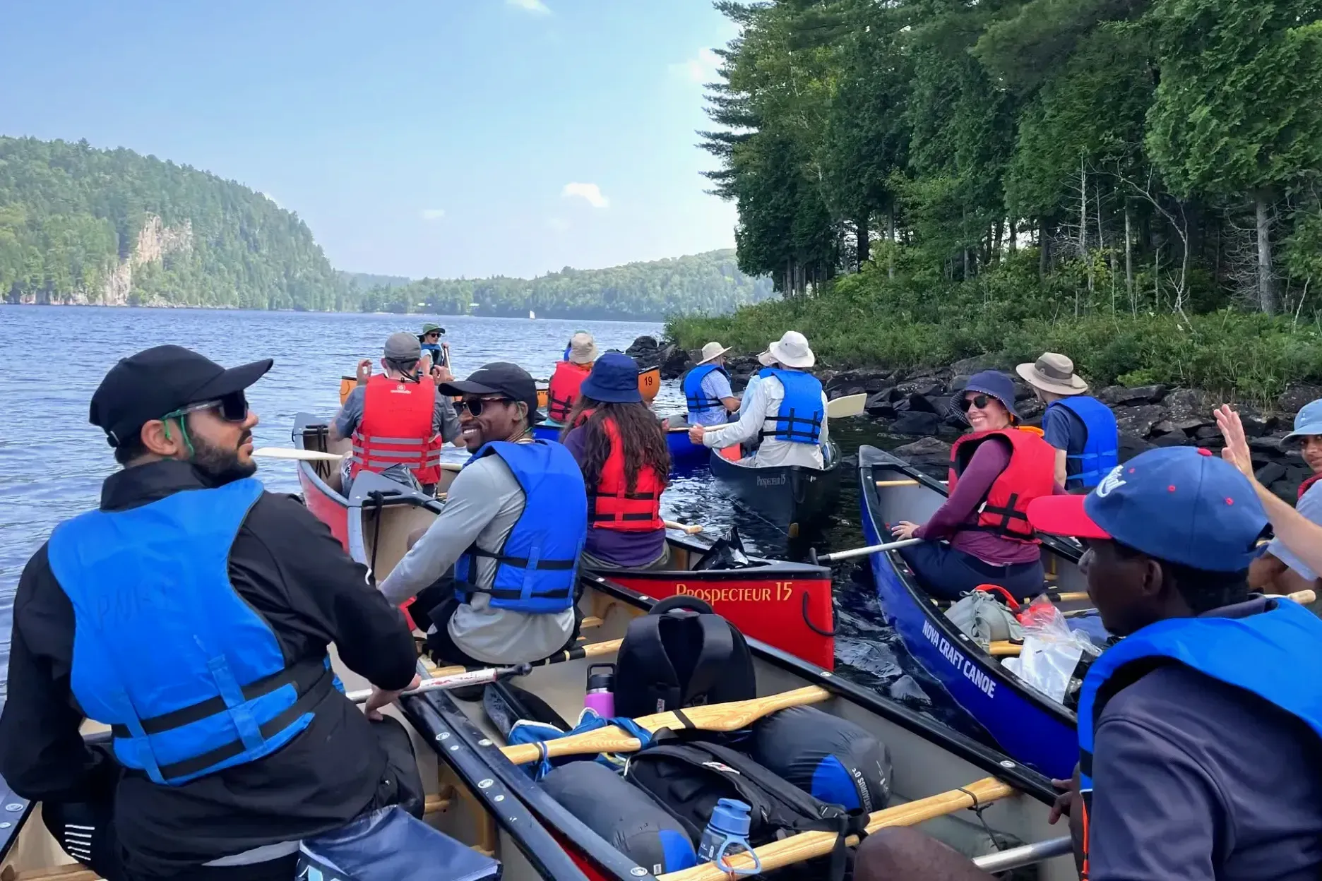 Plusieurs personnes sont installées dans des canots remplis d'équipement de camping, sur un lac, prêtes à partir à l'aventure.