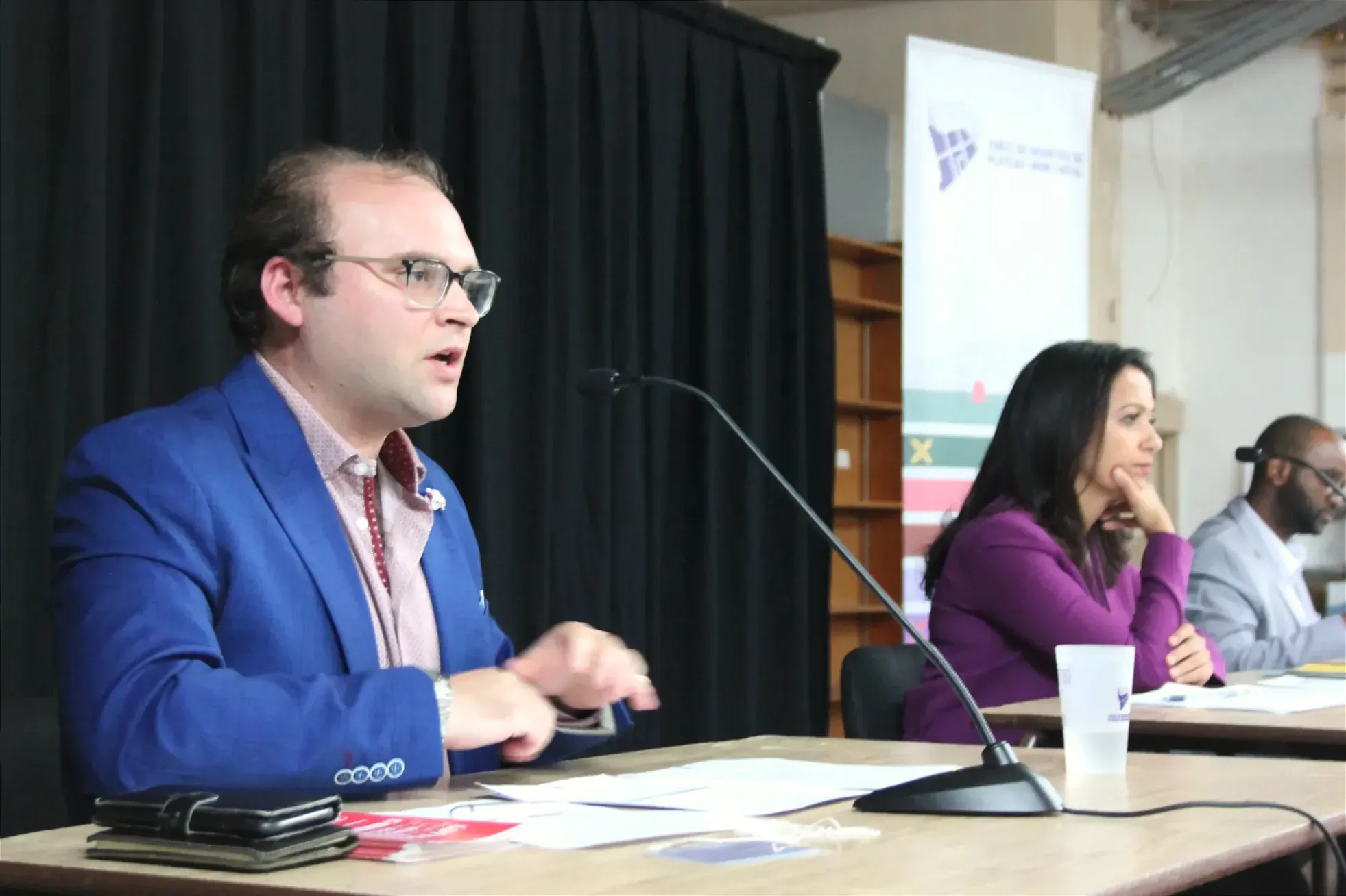 Adrien Welsh prend la parole de son pupitre sur scène. On perçoit derrière lui, à droite, Ruba Ghazal et Thierno Diallo.