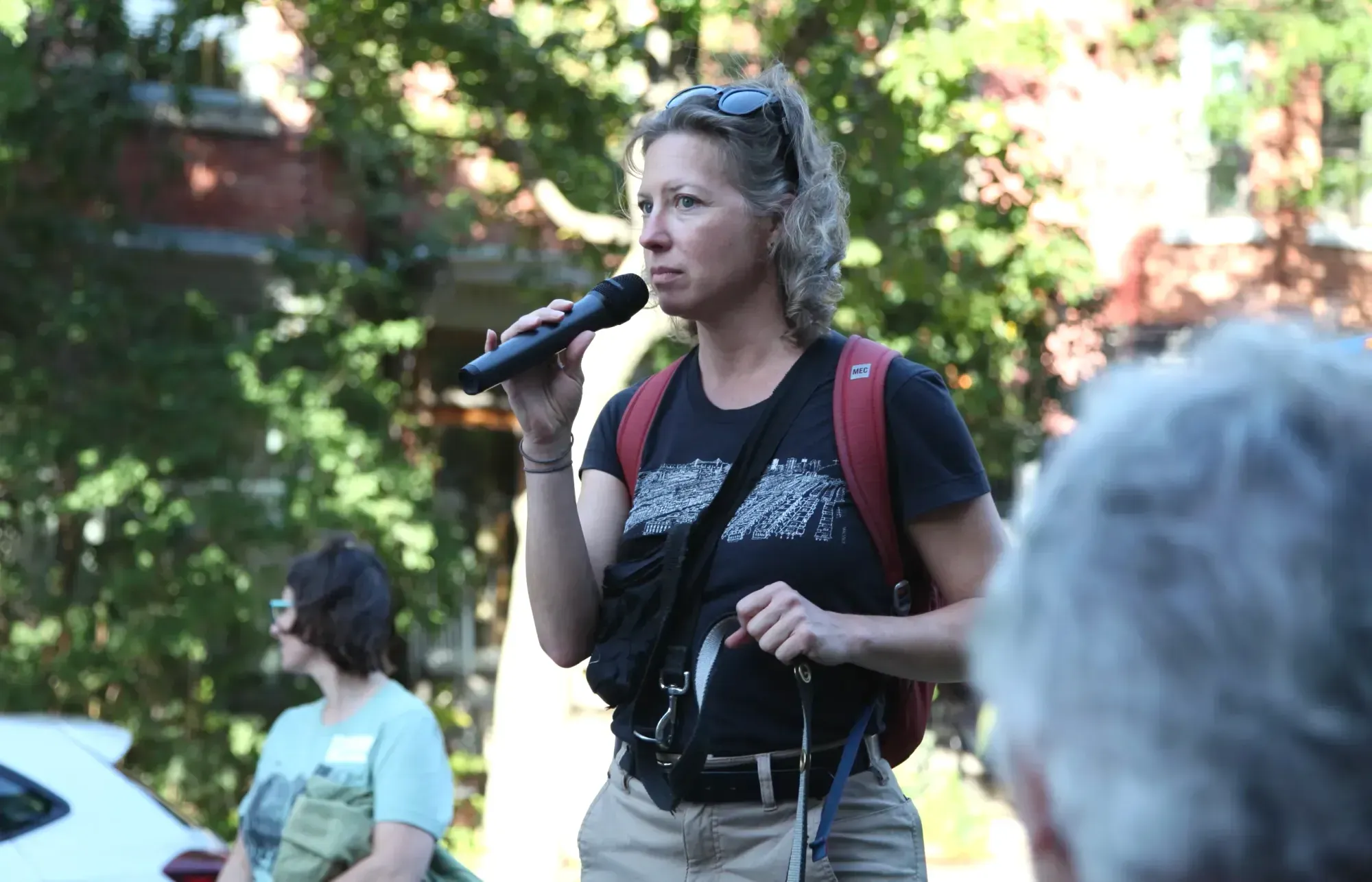 Une dame, l'air préoccupé, tient un microphone devant sa bouche, devant un arrière-plan de murs de brique recouverts de vignes.