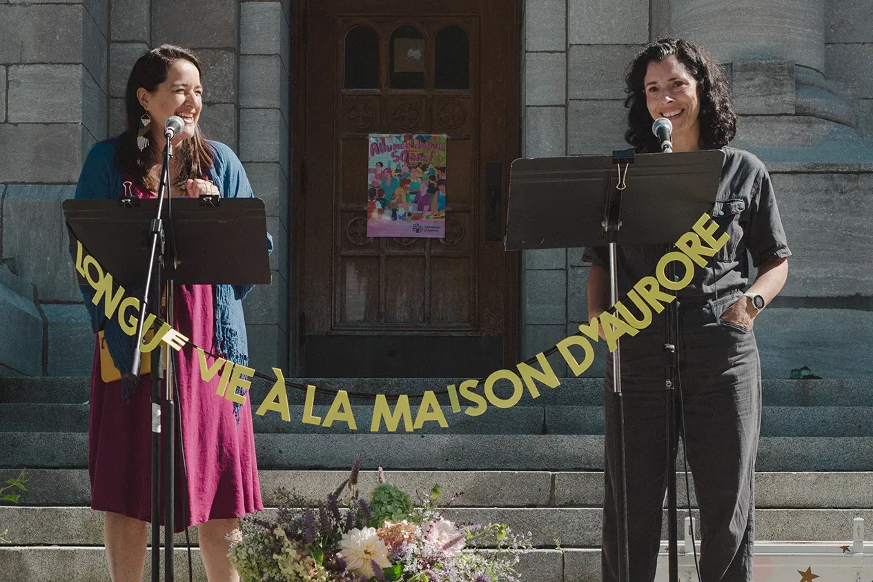 Les deux femmes se tiennent sur le parvis de l'église Saint-Stanislas-de-Kostka, devant deux lutrins décorés d'une banderole où l'on peut lire "LONGUE VIE À LA MAISON D'AURORE"