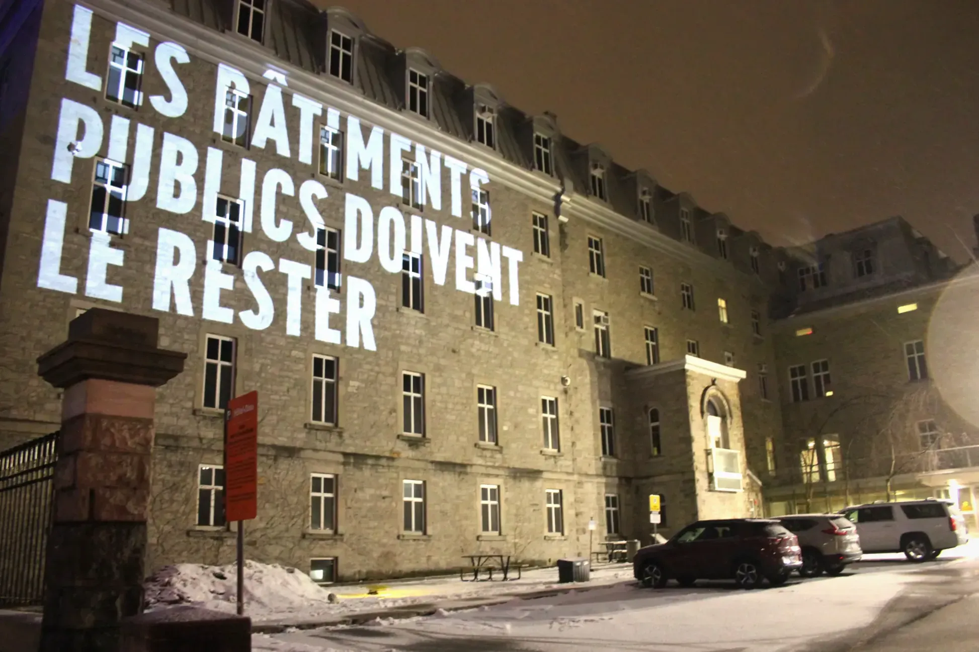 «LES BÂTIMENTS PUBLICS DOIVENT LE RESTER» est projeté sur la façade de l'Hôtel-Dieu de Montréal, lors d'une nuit d'hiver.