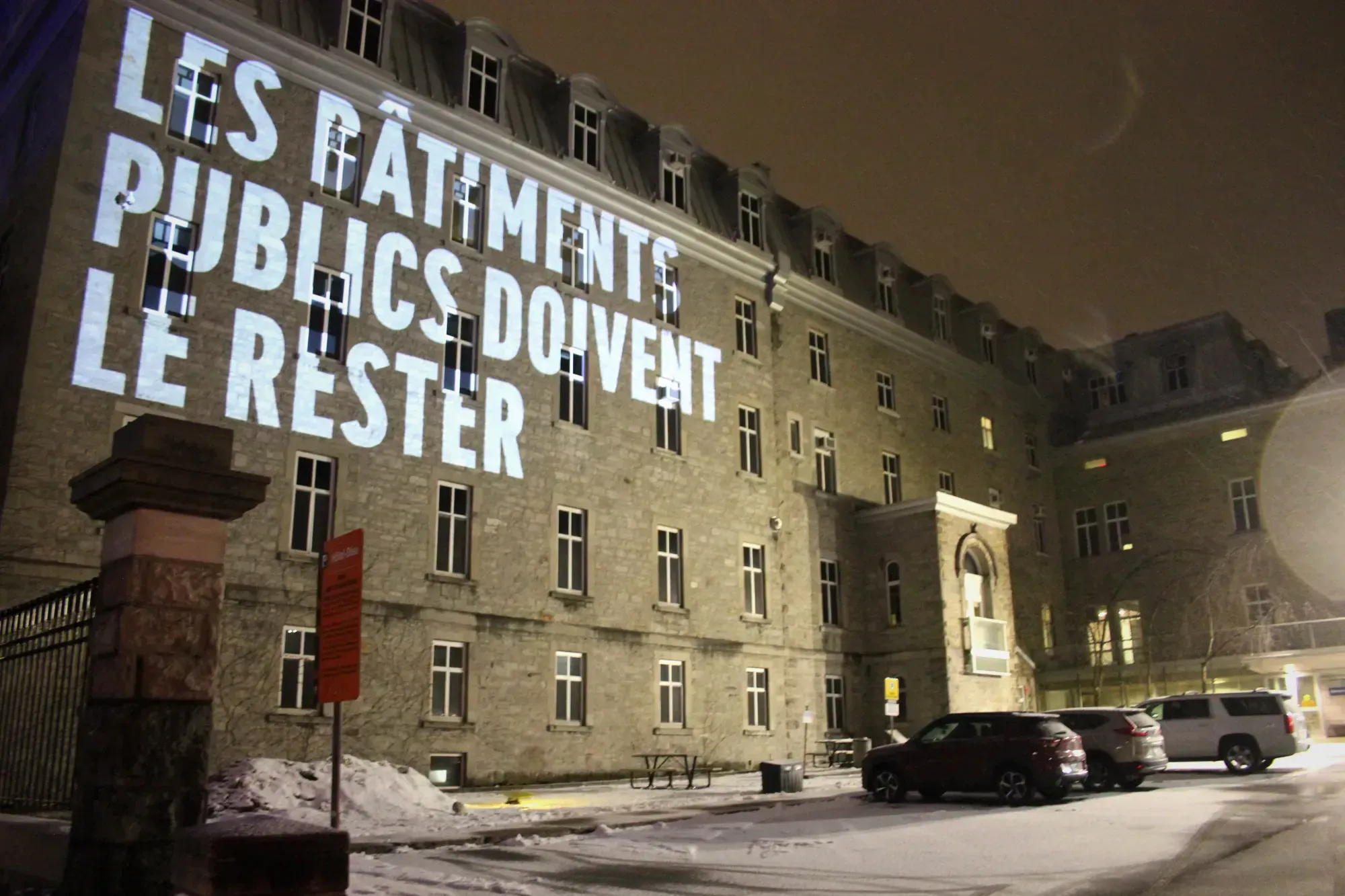 «LES BÂTIMENTS PUBLICS DOIVENT LE RESTER» est projeté sur la façade de l'Hôtel-Dieu de Montréal, lors d'une nuit d'hiver.