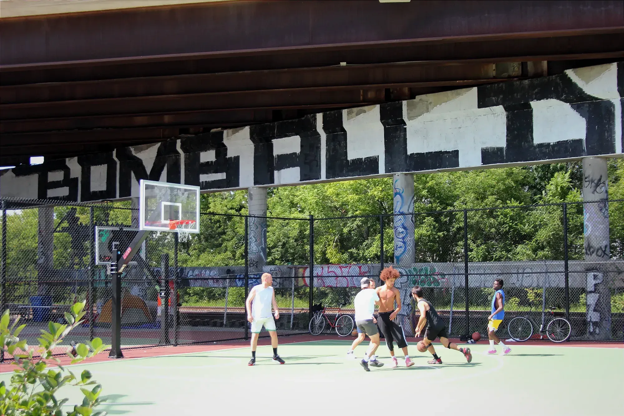 Six hommes jouent en plein soleil sur le sol vert du terrain de basketball extérieur du Mile End. On aperçoit la structure du viaduc Rosemont-Van Horne derrière et au-dessus d'eux. 