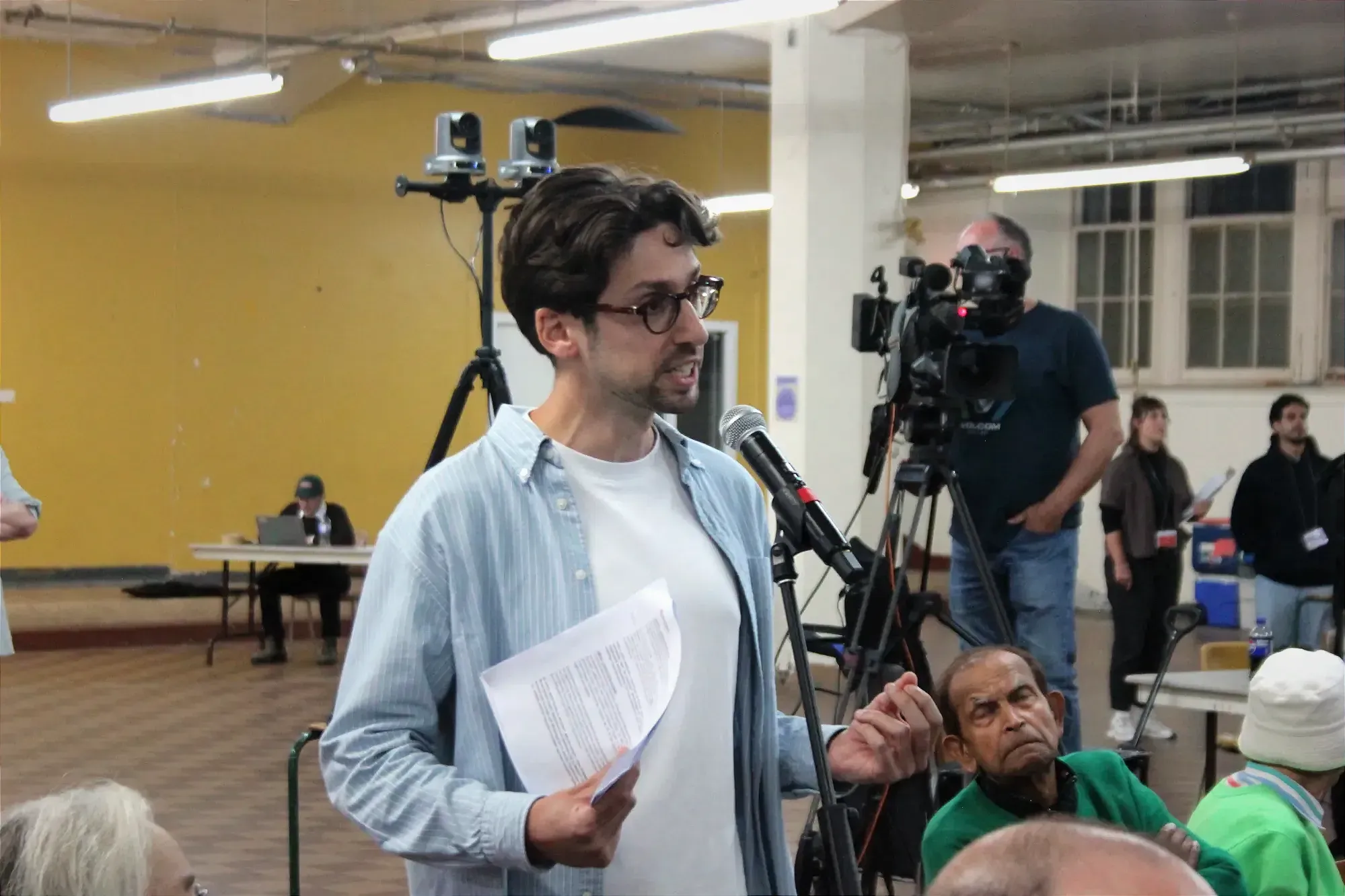 Maxime Claverie prend la parole au micro dans une allée entre des places assises et devant un mur jaune-orange.