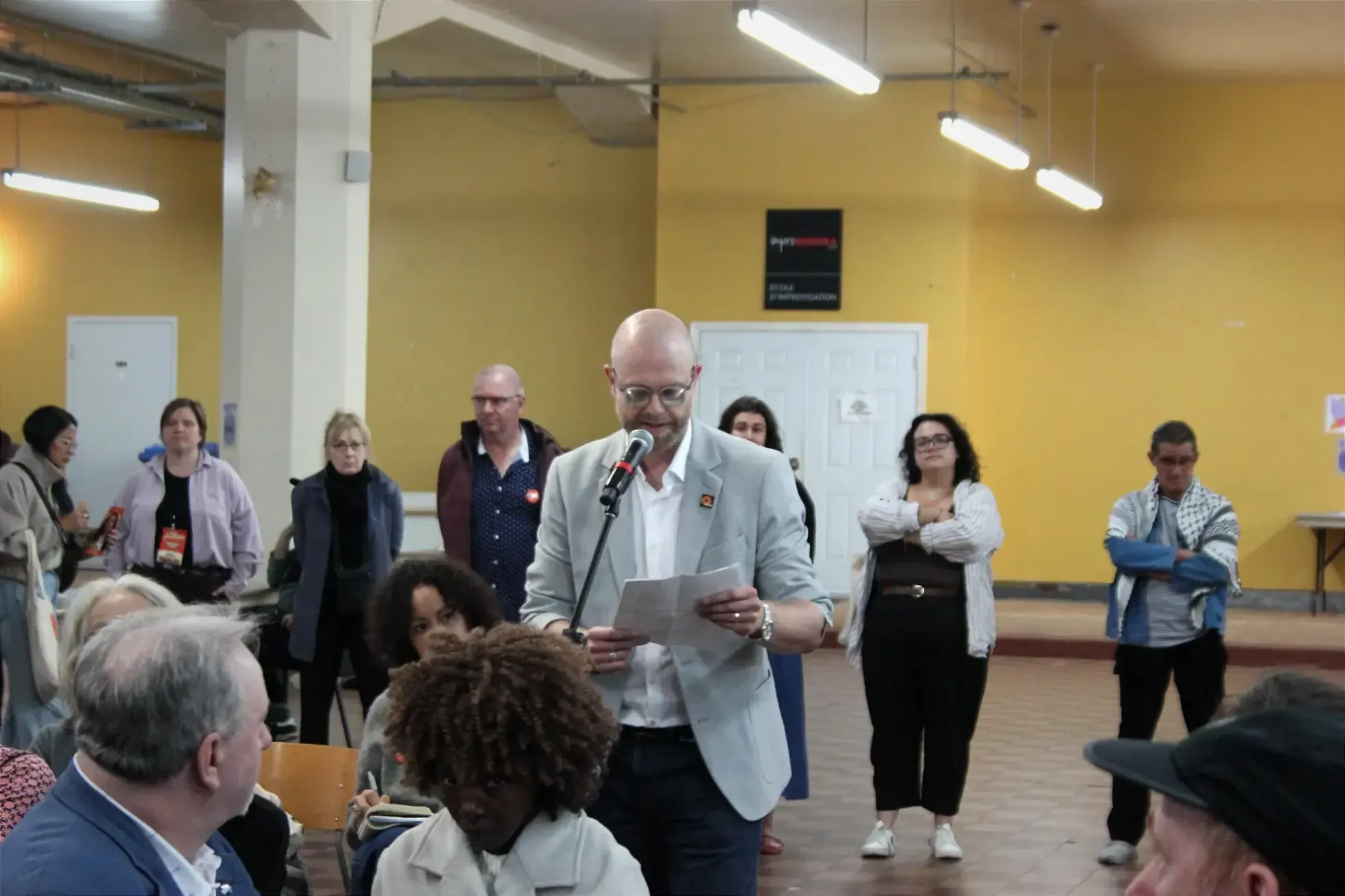 Nicolas Quirion se tient debout devant un micro placé derrière le public assis et lit à partir d'une feuille qu'il tient dans ses maines.