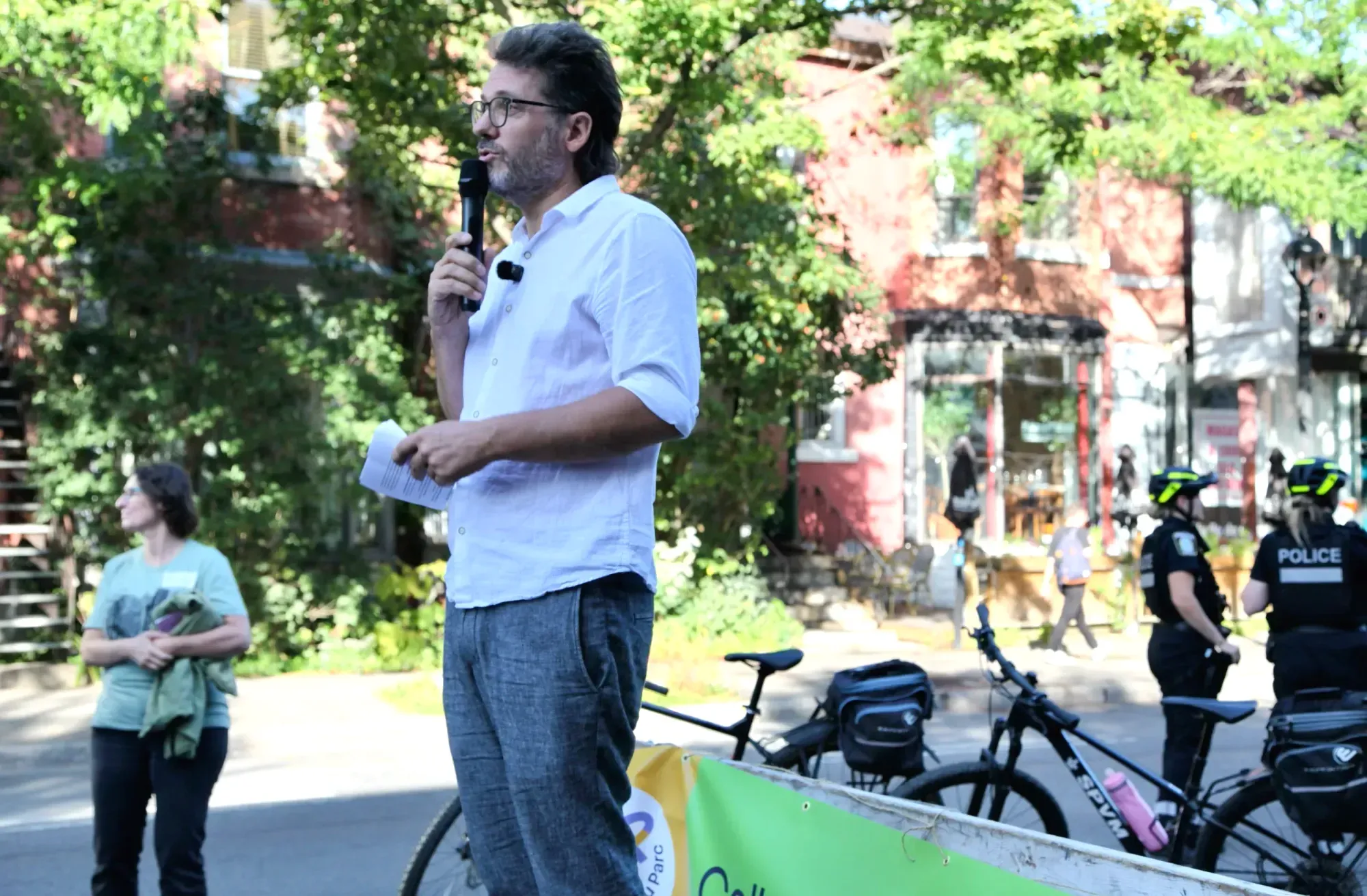 Yan Kestens prend la parole (et le micro) sur l'avenue du Parc. On voit des bâtisses derrière lui, ainsi que deux policières avec leurs casques de vélo sur la tête.