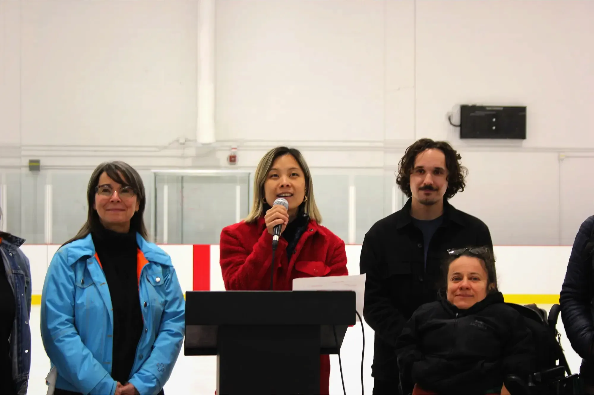 Cathy Wong prend un microphone et la parole, derrière un podium, sur la patinoire intérieure, entourée de Marie Plourde, Gabriel Fortin et Laurence Parent.