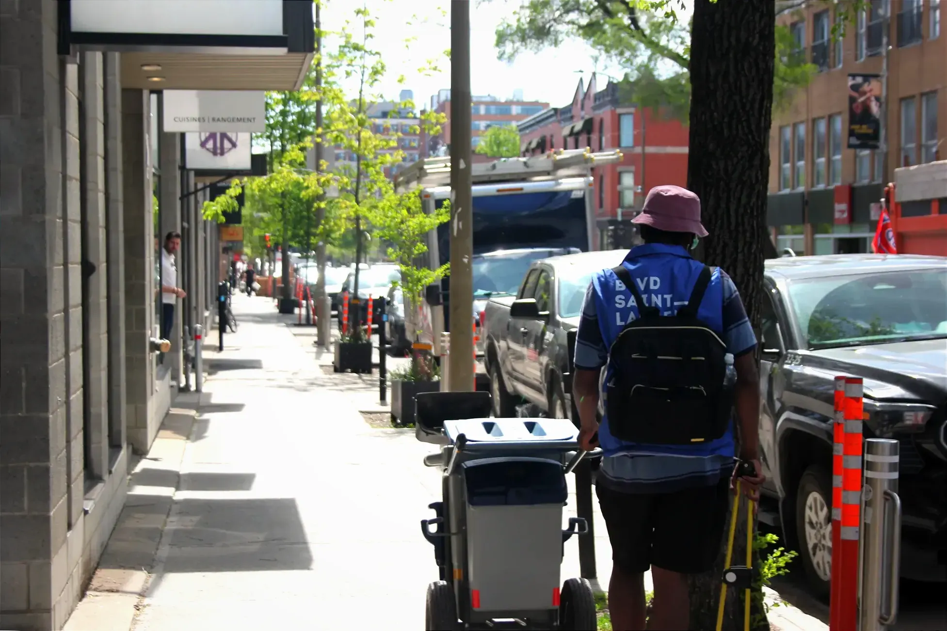 Un homme poussant un chariot d'entretien sur le trottoir du boulevard Saint-Laurent porte une veste bleue et semble ramasser des déchets avec une pince.